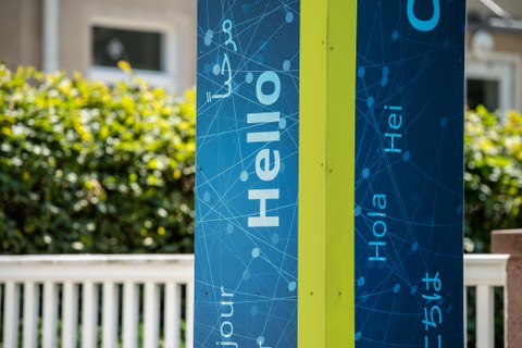 Foto einer Säule in TUD-Blau und Grün mit den Worten "Hello", "Hola", "Hei" und weiteren Sprachen