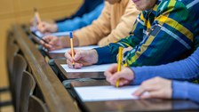 Bild zeigt fünf Studierende, die nebeneinander im Hörsaal sitzen und eine Prüfung schreiben.