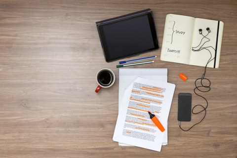 Das Foto zeigt mehrere Gegenstände auf einem Tisch. Man erkennt ein Tablet, mehrere Stifte, einige Zettel, ein Notizbuch, ein Handy und eine Tasse Kaffee
