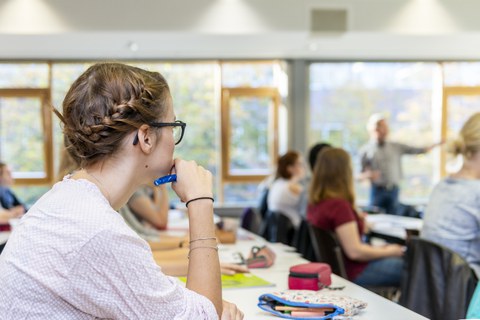 Mehrere Studenten sitzen in einer Vorlesung und schauen zum Dozenten, der gerade etwas erklärt