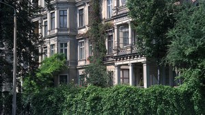 Foto eines mehrstöckigen Altbaus. Teile des Altbaues werden von einer grünen Hecke einem Baum verdeckt. An der Fassade ranken Grünpflanzen empor.