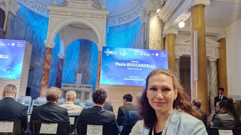 Prof.in Oksana Makovoz auf der Konferenz in Rom. Im Hintergrund sind der prachtvolle Raum, die Teilnehmenden und eine PowerPointPräsentation zu erkennen