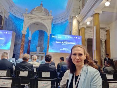 Prof.in Oksana Makovoz auf der Konferenz in Rom. Im Hintergrund sind der prachtvolle Raum, die Teilnehmenden und eine PowerPointPräsentation zu erkennen