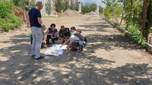 Students in the middle of the picture looking at a map. The place is surrounded by trees. In the background is a row of houses.