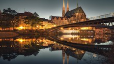 Blick über die Neisse auf die Altstadtbrücke und die abendlich erleuchtete Görlitzer Altstadt mit der prägnanten Silhouette der Peterskirche. Die beleuchtete Altstadt spiegelt sich im Fluss.