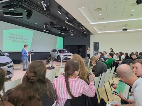 Teilnehmerinnen und Teilnehmer des 2. ACC-Forums während des Besuchs des Skoda-Trainingszentrums in Mlada Boleslav. Im Hintergrund sind der Referent und neue Fahrzeuge zu erkennen.