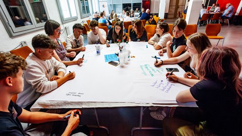 Schülerinnen und Schüler sitzen um einen Tisch, auf dem Workshop-Materialien verteilt sind.