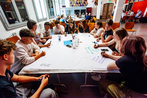 Schülerinnen und Schüler sitzen um einen Tisch, auf dem Workshop-Materialien verteilt sind.