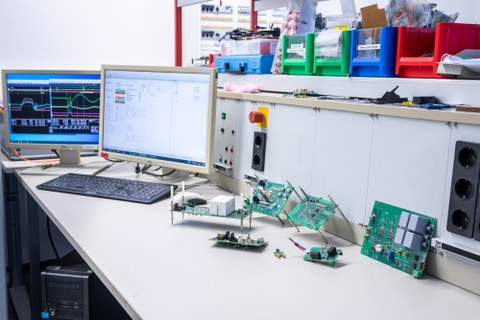 Im Vordergrund: Leiterplatten (PCBs) zur Charakterisierung von GaN Transistoren und Erprobung unterschiedlicher Ansteuer- und Schutzkonzepte. Im Hintergrund: PC-Arbeitsplatz zum Teststand.