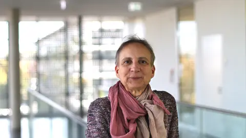 Anja Hampel im Treppenhaus des Werner-Hartmann Baus schaut freundlich in die Kamera. Sie trägt einen rot-weiß-schwarz melierten Pullover und ein hellrotes Tuch.