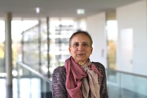 Anja Hampel im Treppenhaus des Werner-Hartmann Baus schaut freundlich in die Kamera. Sie trägt einen rot-weiß-schwarz melierten Pullover und ein hellrotes Tuch.