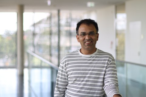 Rabiul Islam wears a horizontall striped pullover with a black and white stripes. He wears glasses and ssmiles towards the camera.