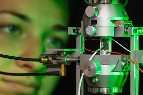 Ein Messsensor mit gelöteten Kontakten eingebaut in einer Halterung zum charakterisieren des Sensors. Grünes Licht strahlt die Probenhalterung von rechts an. Im Hintergrund ist unscharf das Gesicht einer Person zu erkennen, welche den Sensor anschaut.
