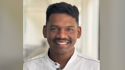 Sudhi is smiling at the camera in a softly lit indoor hallway, wearing a white collared shirt.