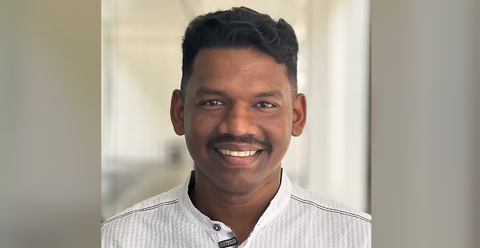 Sudhi is smiling at the camera in a softly lit indoor hallway, wearing a white collared shirt.