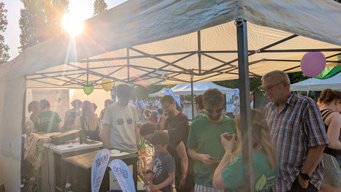 Mehrere Menschen stehen im Zelt vom Excellenz-Cluster REC². Der Versuchsuafbau zu leuchtenden Drinks und der Aufbau zur Zitronenbetriebenen Funkempfänger werden gezeigt.