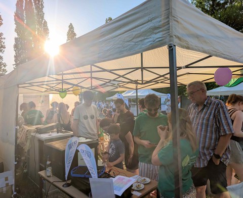 Mehrere Menschen stehen im Zelt vom Excellenz-Cluster REC². Der Versuchsuafbau zu leuchtenden Drinks und der Aufbau zur Zitronenbetriebenen Funkempfänger werden gezeigt.