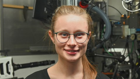 Frau Miriam Heise im Labor vor einer Anlage und lächelt in die Kamera. Sie trägt ein schwarzes Oberteil, Brille und hat blonde Haare. 