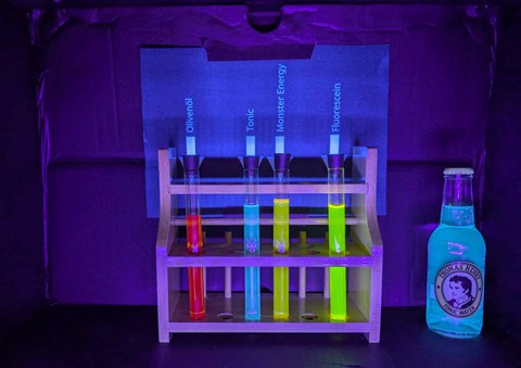 Die drei Haushaltsüblichen Stoffe Olivenöl, Tonic Water und ein Energy Drink sowie Fluorescein werden mit UV Licht angeleuchtet. Dadurch leuchten die flüssigkeiten in den Reagenzgläsern Rot, Blau, Gelb und Grün auf.