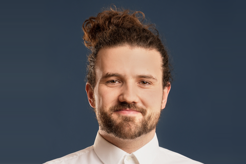 The person Ron Hildebrandt in a white suit top and a soft smile looking directly into the camera. He has brown eyes, long brown pinned up hair and wears a beard. The background is dark blue. 