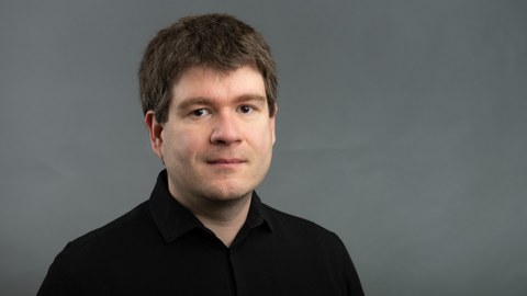 Christain Ullmann in schwarzen Hemd schaut direkt in die Kamera mit leichtem lächeln. Der Hintergrund ist neutral grau.