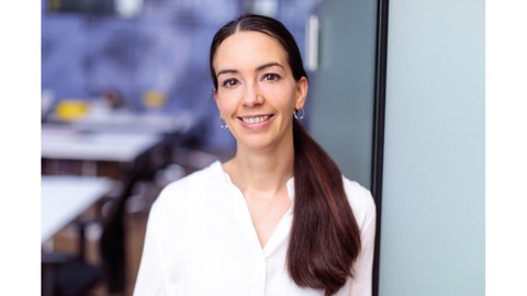 Professor Elizabeth von Hauff trägt ein weißes Hemd und lächelt glücklich in die Kamera. Im Hintergrund ist ein unscharfer Besprechungsraum zu sehen.