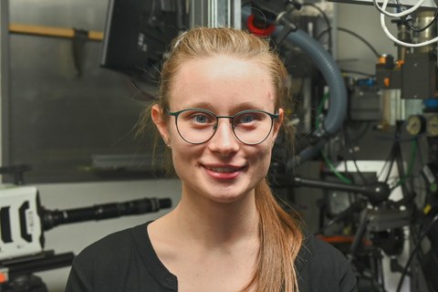 Frau Miriam Heise im Labor vor einer Anlage und lächelt in die Kamera. Sie trägt ein schwarzes Oberteil, Brille und hat blonde Haare. 