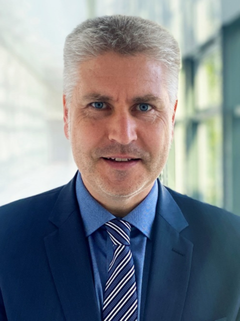 Prof. Härtling trägt ein dunkelblauen Anzug mit Hellblauen Hemd und schräg gestreifer Krawatte.