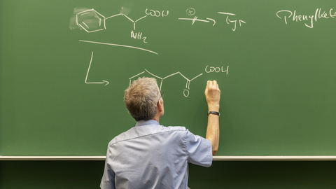A green chalkboard with chemical formulas of organic substances that are being written on by a professor wearing a blue shirt.