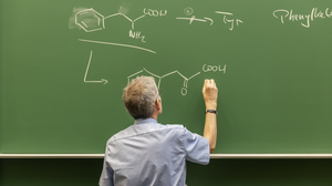 A green chalkboard with chemical formulas of organic substances that are being written on by a professor wearing a blue shirt.