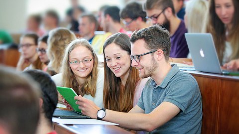 Bild: junge Menschen sitzen im Hörsaal, lächeln und schauen sich gemeinsam Unterlagen an