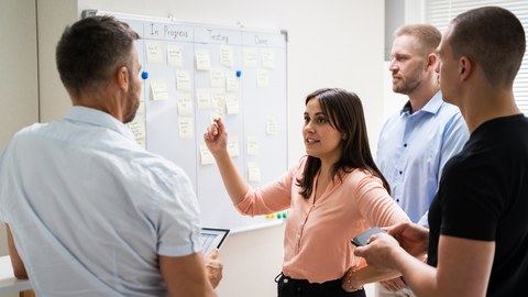 Vier Personen stehen vor Whiteboard mit Notizzetteln, eine Person erklärt etwas.