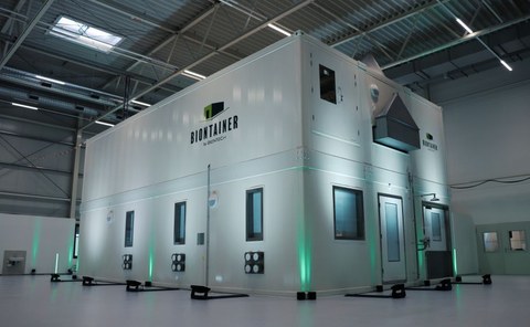 White containers form a modular process plant. This stands in a hall and is partially illuminated in green.