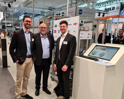 Drei Personen in schicker Kleidung auf einem Stand auf der Hannover Messe. Im Hintergrund ist eine modulare Prozessanlage zu sehen.