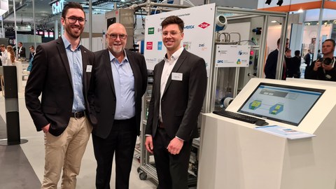 Three people in smart clothes at a booth at the Hannover Messe. A modular process plant can be seen in the background.