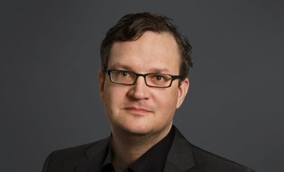 Man with short dark hair and glasses, dressed in dark suit and shirt