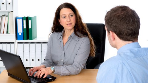 Auf dem Foto ist eine Frau am Schreibtisch an Ihrem PC zu sehen. Sie hat die Hände auf der Tastatur und unterhält sich mit einem Mann, der vor ihr sitzt.