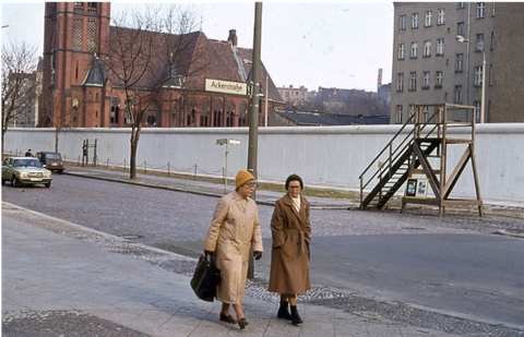 Foto. Zwei ältere Damen in braunen Mänteln mit gestrickten Mützen laufen einen Gehweg entlang. Direkt hinter ihnen ein Straßename, "Ackerstraße" an einem Laternenpfahl. Auf der anderen Straßenseite verläuft hinter dem Gehweg und einem schmalen Grünstreif