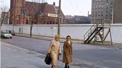 Foto. Zwei ältere Damen in braunen Mänteln mit gestrickten Mützen laufen einen Gehweg entlang. Direkt hinter ihnen ein Straßename, "Ackerstraße" an einem Laternenpfahl. Auf der anderen Straßenseite verläuft hinter dem Gehweg und einem schmalen Grünstreif