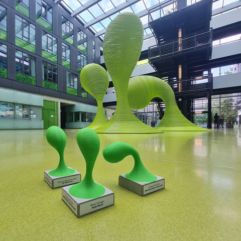 Im Hintergrund sind die tropfenförmigen grünen Stahlskulpturen aus dem Foyer, im Vordergrund sind Miniaturausgaben dieser Skulpturen auf Betonsockeln mit Aufschriften der Preistragenden.