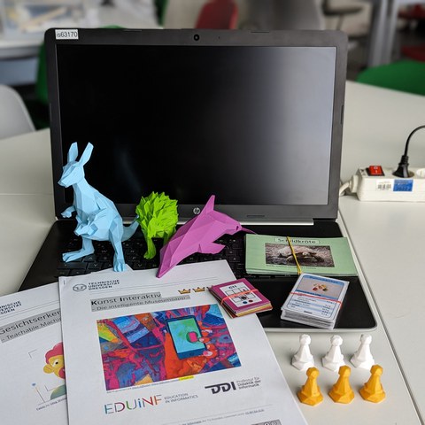 Auf dem Bild sind verschiedene Materialien für den KI-Workshop abgebildet. Auf einem Laptop stehen mehrere 3D-gedruckte Tiere. Daneben liegen anderen Unplugged Materialien und Arbeitsblätter.