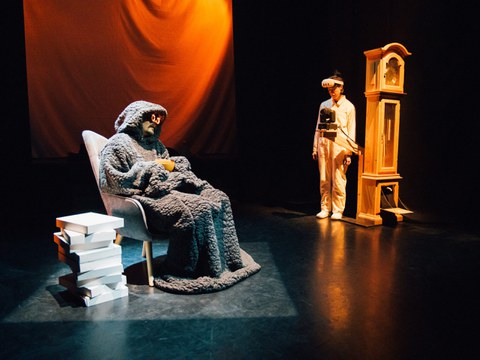 Photo from the theater act: Earl Grey sitting in his chair, talking to Jeannie next to a clock.