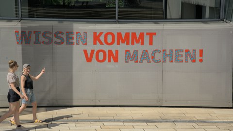 Foto von zwei Personen, die über den Campus laufen. Eine davon zeigt auf das Banner "Wissen kommt von machen!"