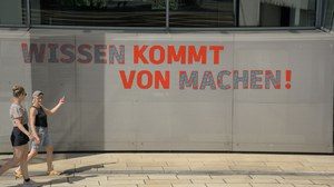 Foto von zwei Personen, die über den Campus laufen. Eine davon zeigt auf das Banner "Wissen kommt von machen!"