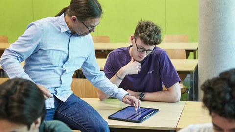 Situation während einer Lehrveranstaltung - Lehrender am Platz eines Studierenden im Gespräch mit einem Tablett auf dem Tisch. 