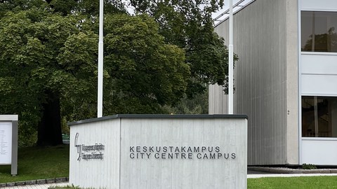 Im Vordergrund grüne Wiese, im Hintergrund das Zeichen der University of Tampere