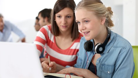 The photo shows two female students looking at a laptop together and taking notes in a notebook.