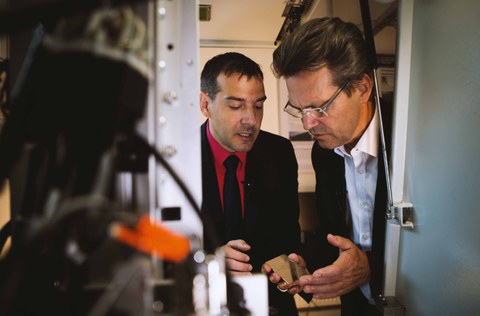 Professor Mücklich rechts hält ein laserstrukturiertes Edelstahlsubstrat in der Hand und Professor Lasagni links erklärt wie es strukturiert wurde 
