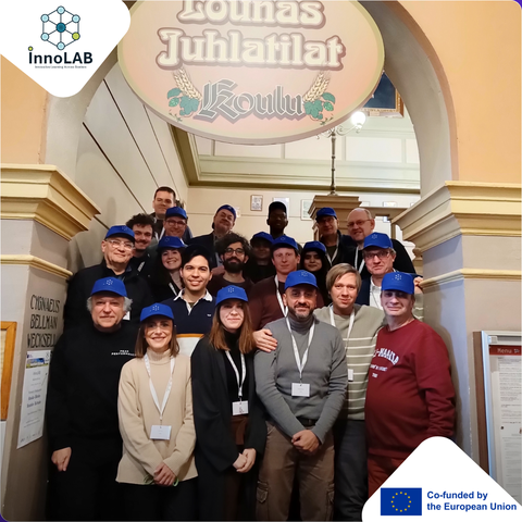 Eine Gruppe von etwa zwanzig Personen mit passenden blauen Caps steht gemeinsam auf einer Innentreppe unter einem dekorativen Schild. Die meisten lächeln und tragen Namensschilder. 