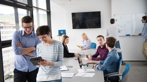 Foto von acht Menschen in einem Büro, zwei von ihnen sind im Vordergrund und schauen auf ein Tablet. Die anderen sitzen sprechend an einem Tisch beziehungsweise stehen vor einem Whiteboard.